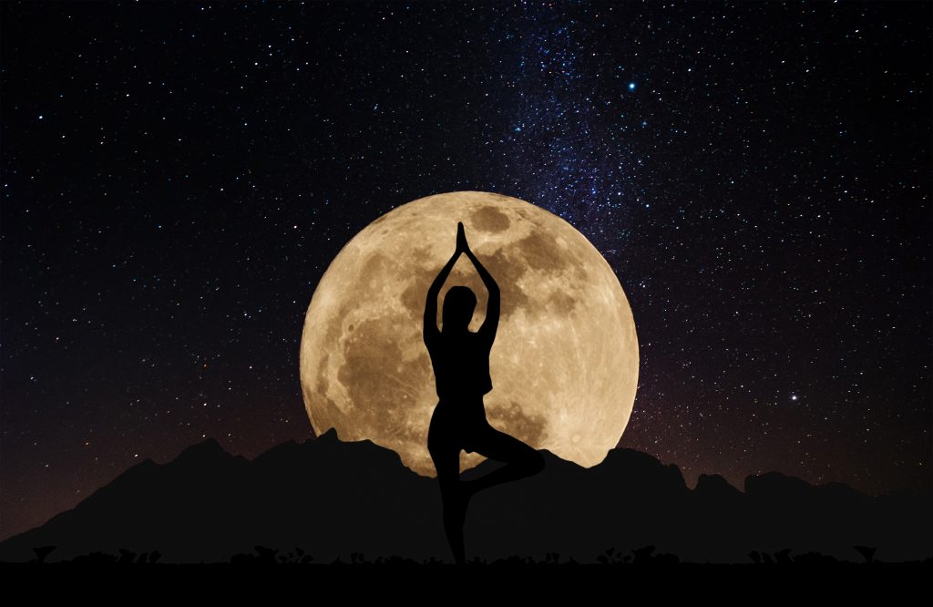 Silhouette young woman practicing yoga pose at night under full moon with sky full of stars